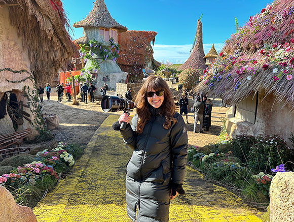 A woman in a black puffer coat holds a large camera in a village with whimsical, thatched-roof cottages.