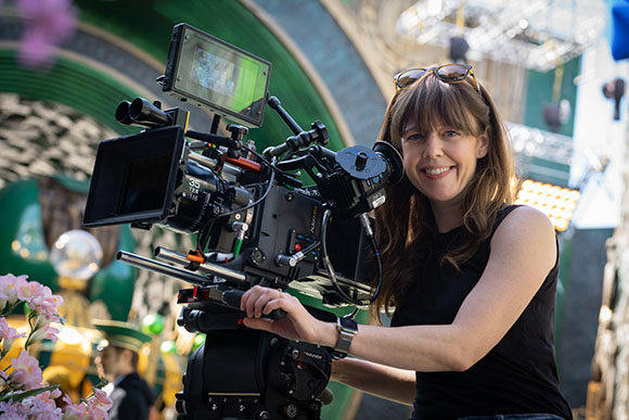 A smiling woman with long brown hair stands behind a large professional cinema camera on a film set.