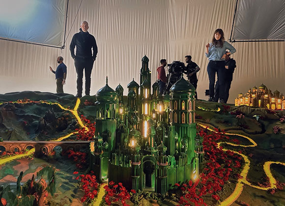 Film crew stands around a large, illuminated green miniature castle model set within a landscape.
