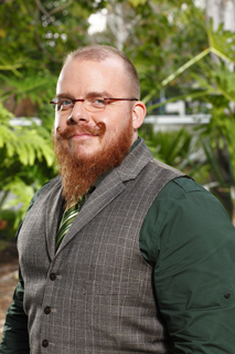 A man with a long red beard, glasses, a grey patterned vest, and a green shirt stands outdoors.
