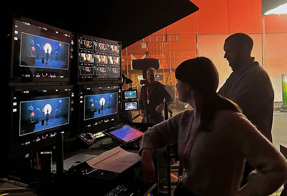 Film crew members stand in a dark studio looking at multiple glowing production monitors.