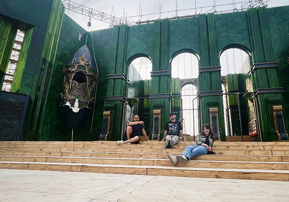 Three people sit on wide wooden steps before a massive, dark green theatrical set with arched windows.