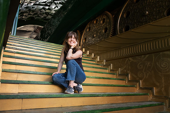A smiling woman in a black tank top and jeans sits on wide, golden-toned stairs near large decorative gears.