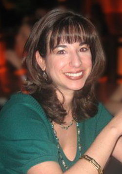 A smiling woman with dark hair and bangs wearing a green top and jewelry.