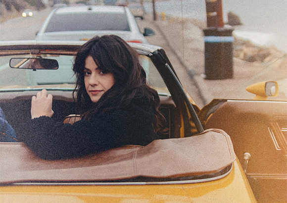 A woman with long dark hair looks over her shoulder while sitting in a yellow convertible car.
