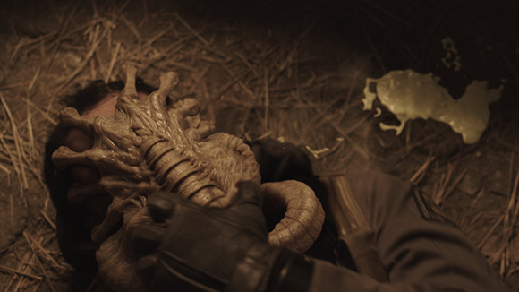 A textured, alien-like creature head presses against a person lying on straw.