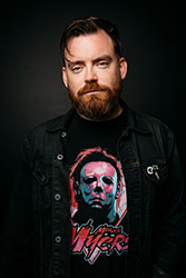 A man with a reddish-brown beard and dark hair wearing a black denim jacket over a Michael Myers graphic t-shirt.