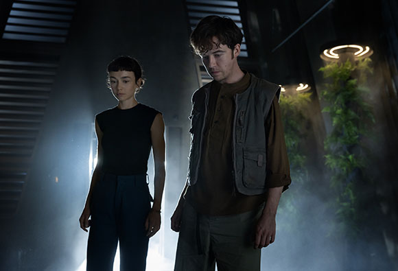 A woman in a black sleeveless top and a man in a brown vest walk through a misty, dark hallway.