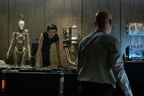 A person leans over a table of equipment in a dark lab near a skeleton and a man in a light shirt.