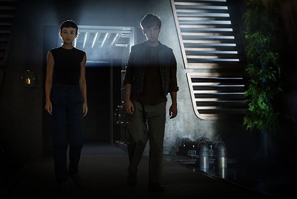 A woman in a black sleeveless top and a man in a brown jacket walk through a dark, industrial hallway.