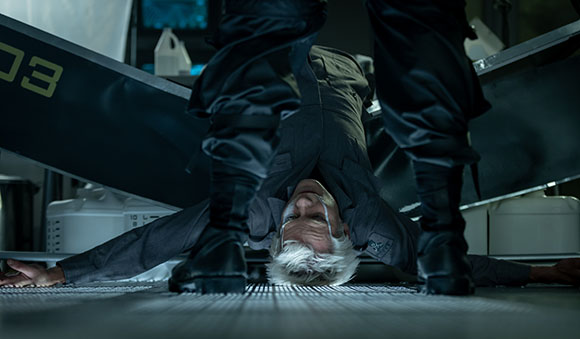 A man with white hair lies on a metal floor while another person's legs stand over him.