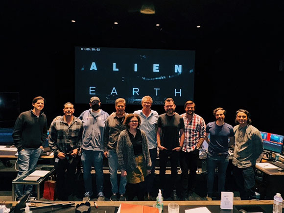 Ten people standing in a row in front of a large screen that reads 'ALIEN EARTH' in white capital letters.