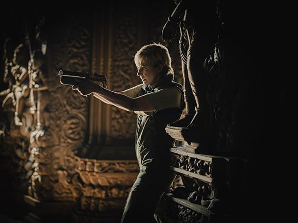 A woman in green overalls aims a handgun while standing near a large stone statue in a dark, carved room.