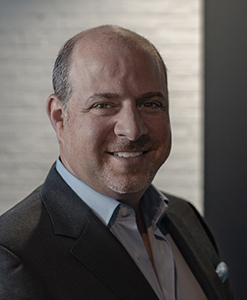 A smiling man with short hair and a goatee wearing a dark blazer and light blue button-down shirt.