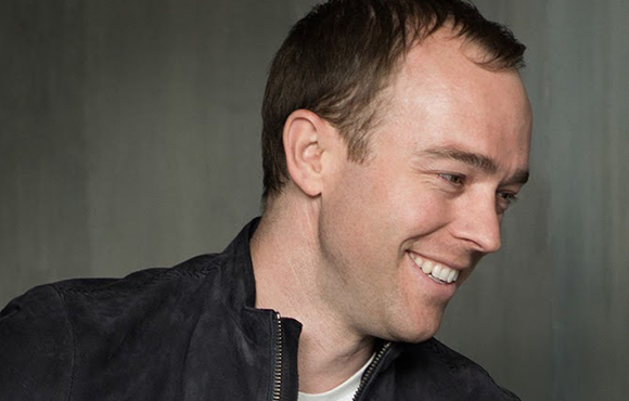 A man with short brown hair smiling while looking to the side against a gray background.