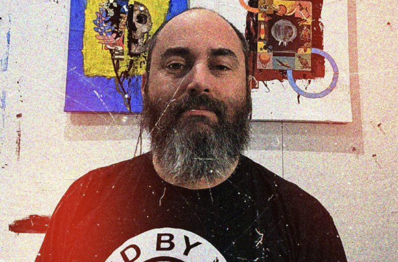 A man with a thick dark beard and balding head looks at the camera in front of abstract paintings.
