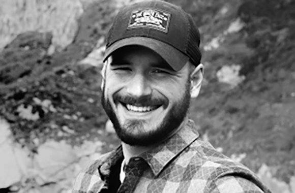 A bearded man in a baseball cap and plaid shirt smiles outdoors.