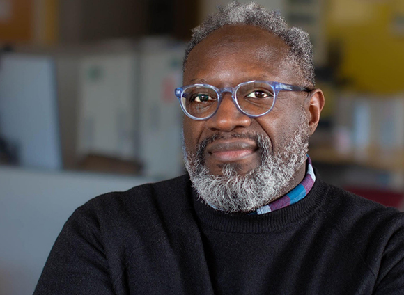 A man with graying hair and a beard wearing blue-rimmed glasses and a black sweater.
