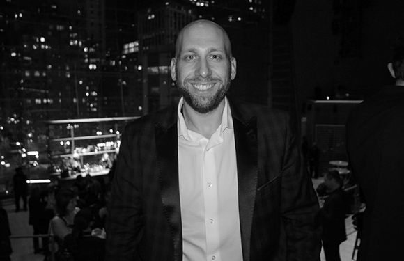 A smiling man in a dark blazer and white button-down shirt at an outdoor nighttime event.