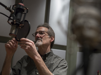 A man with glasses and a goatee adjusts a large studio microphone.