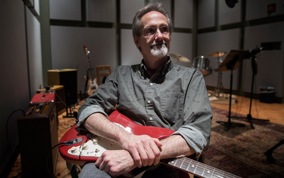 A man with glasses and a grey beard sits in a music studio holding a red electric guitar.