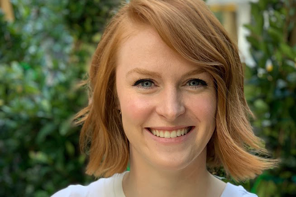 A smiling woman with shoulder-length reddish-blonde hair and blue eyes against a blurred green background.