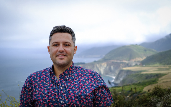 A man in a patterned blue shirt smiling against a backdrop of misty coastal cliffs and the ocean.