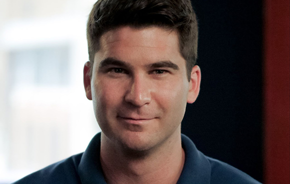 A close-up portrait of a man with dark hair smiling slightly against a blurred background.