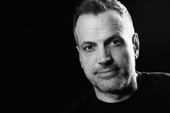 Black and white close-up portrait of a man with short hair and light stubble looking toward the camera.