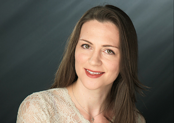 A smiling woman with long brown hair wearing a lace top and a silver necklace against a dark background.