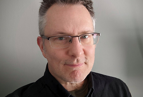 A man with short graying hair and glasses looking toward the camera against a plain wall.