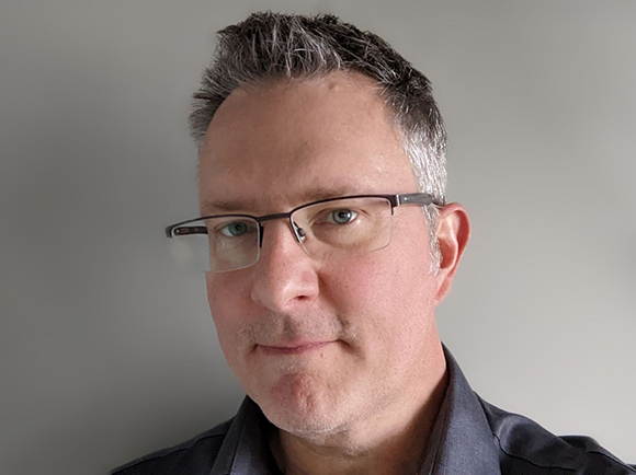 A man with short graying hair and glasses looking forward against a plain gray background.