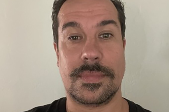 Close-up portrait of a man with dark hair, brown eyes, and a mustache and goatee against a plain wall.