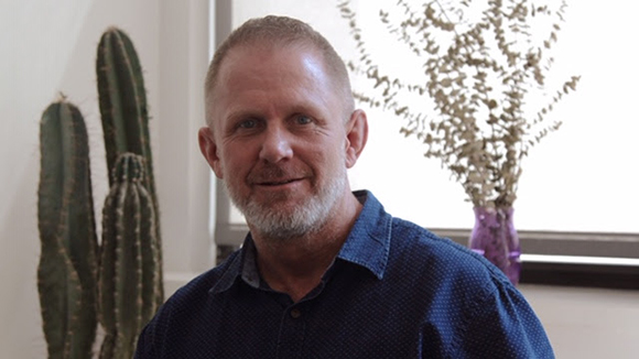 A smiling man with a grey beard wearing a blue button-down shirt.