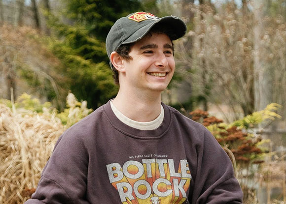 A smiling young man wearing a dark baseball cap and a brown Bottle Rock sweatshirt outdoors.