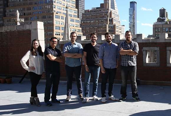 Six diverse adults standing in a row on a sunny rooftop with urban skyscrapers behind them.