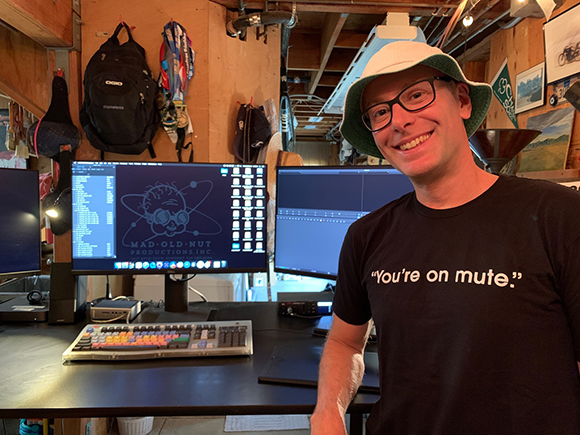 A smiling man in a bucket hat and a black t-shirt that says "You're on mute" stands in a studio.