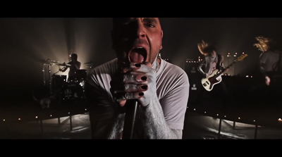 A vocalist sings into a microphone with an open mouth, surrounded by band members in a dark setting.
