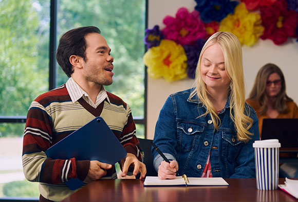 A man and a woman with Down syndrome sit at a table, smiling and looking at a notebook.