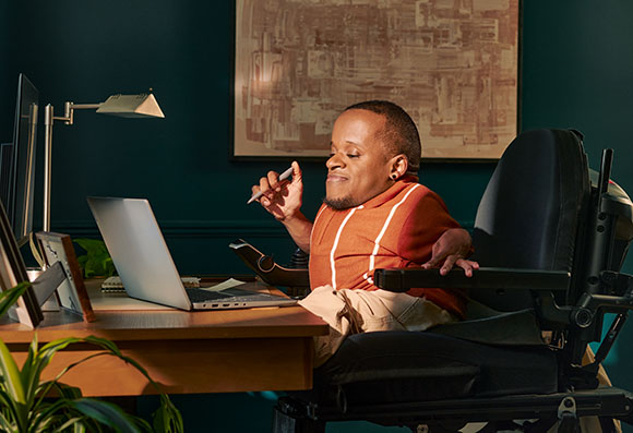 A person in a motorized wheelchair sits at a desk, holding a pen and looking at a laptop screen.