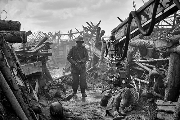 A film camera on a crane overlooks actors lying in a muddy, debris-filled trench.