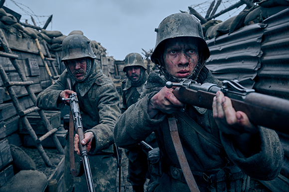 Three soldiers in dirty uniforms and helmets hold rifles in a muddy trench under a cloudy sky.