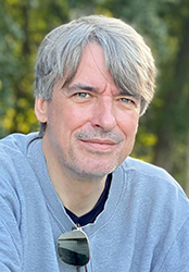 A man with graying hair and a light beard wearing a gray sweatshirt outdoors.