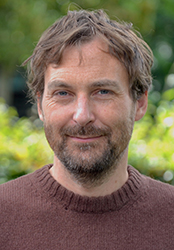 A man with wavy brown hair and a beard wearing a brown sweater, smiling against a blurred green background.
