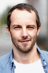 A close-up portrait of a man with short brown hair and a light beard wearing a blue shirt.