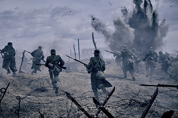 Soldiers in historical military uniforms charge across a muddy battlefield toward a large explosion.