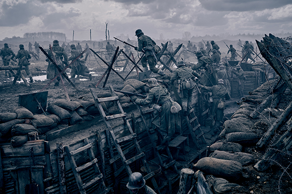 Soldiers in helmets move through a muddy trench filled with sandbags, wooden ladders, and barbed wire.