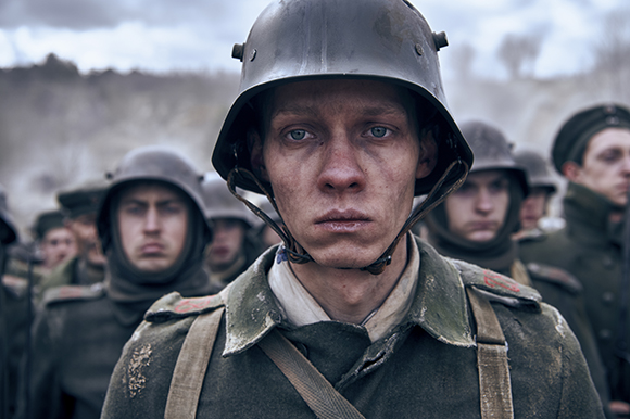 A young soldier with a dirt-streaked face wears a metal helmet and green uniform, surrounded by other soldiers.