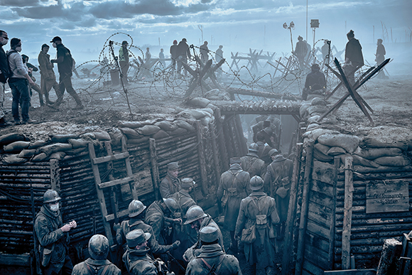 Soldiers in a dirt trench looking toward barbed wire fences and fortifications under a cloudy sky.