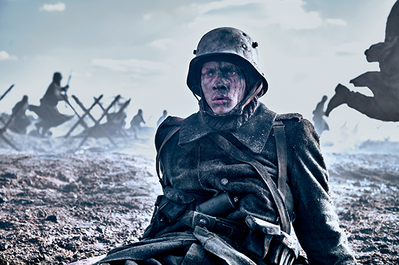 A soldier in a dark uniform and helmet sits in a muddy, war-torn field under a cloudy sky.